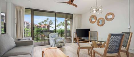 Una sala de estar moderna con sofá, mesa de centro y sillones, televisor y balcón con vista a palmeras.