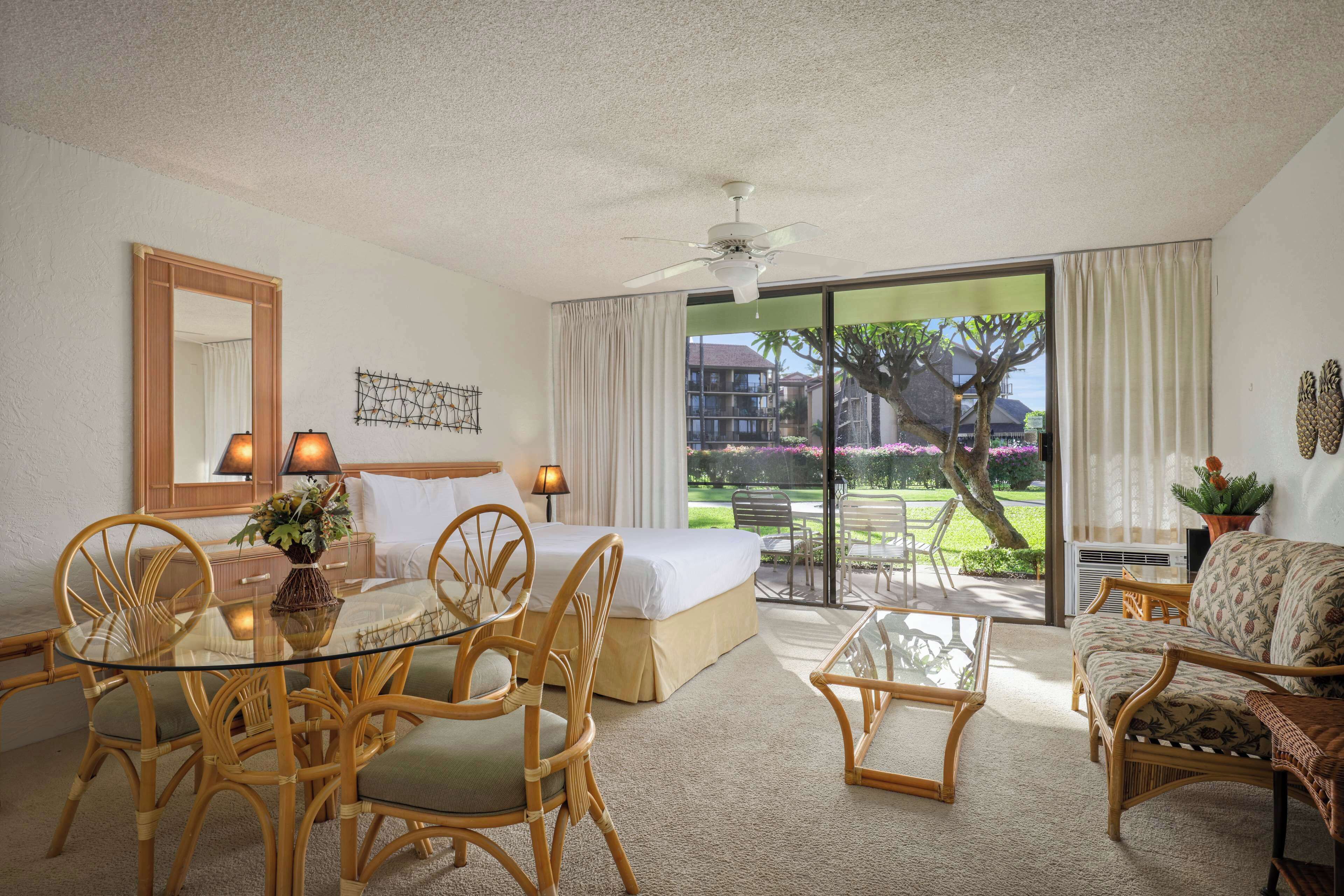 A hotel room with a bed, a glass dining table, wicker chairs, a sofa, a ceiling fan, and a large window with a view of a garden and buildings.