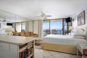 Habitación de hotel con cama, escritorio, silla, estantería, ventilador de techo, televisor y balcón con vista al océano.