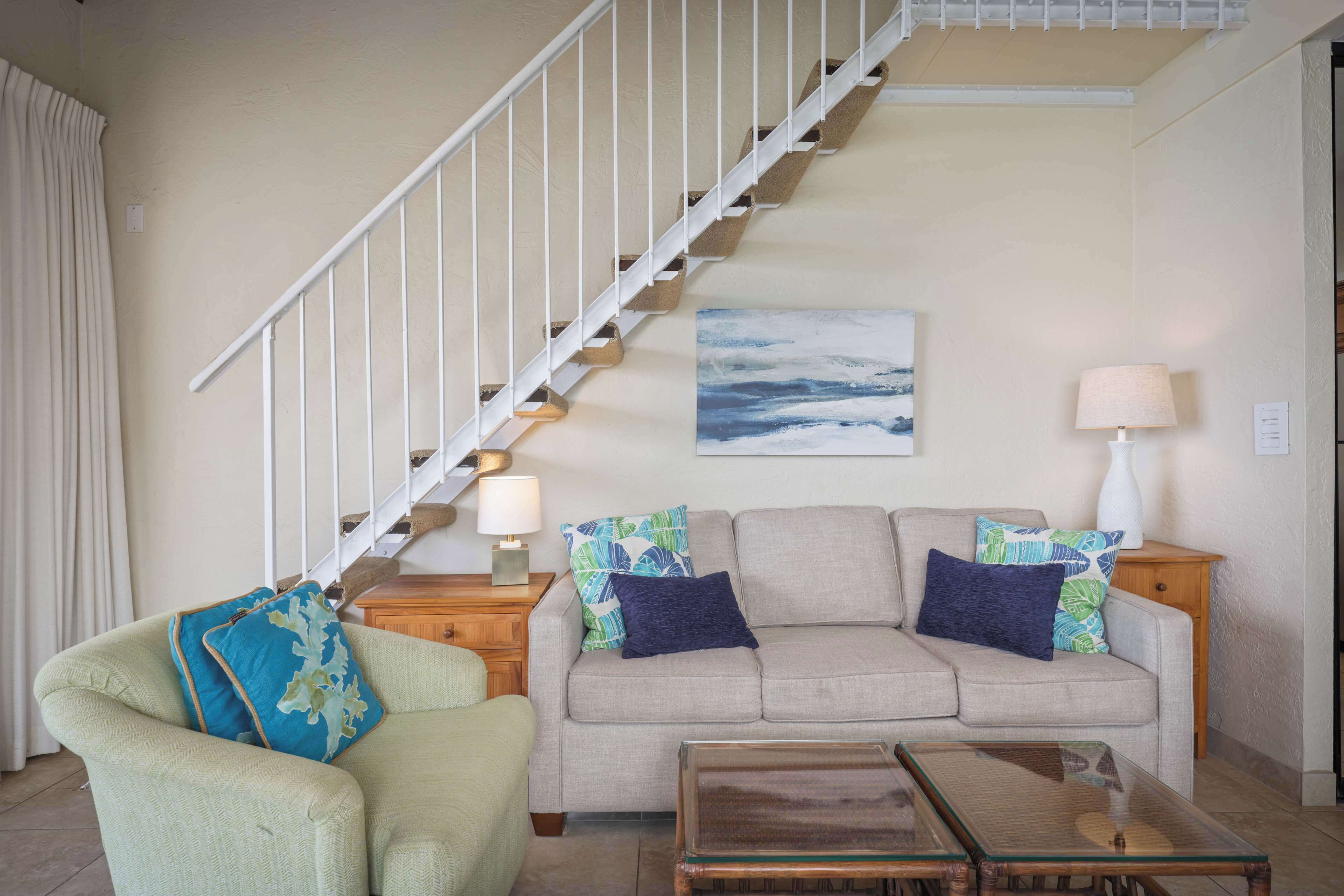 Una sala de estar con una escalera, un sofá color beige, un sillón verde y una mesa de centro de vidrio.