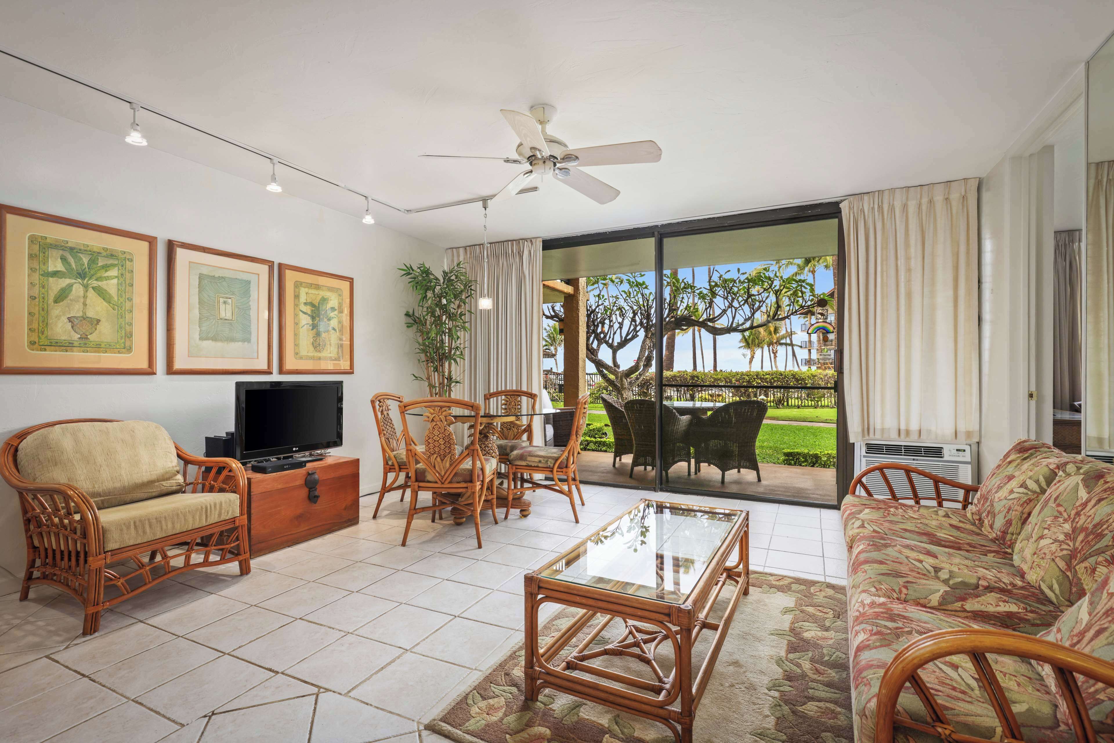 A living room with a TV, wicker furniture, a glass coffee table, and a view of a garden through a sliding door.