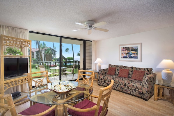 A living room with a glass table, wicker chairs, a sofa, a ceiling fan, and a large window with a view of palm trees and buildings.