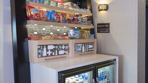 A snack display with various chips and snacks, a sign indicating a breakfast area, and a refrigerator stocked with bottled water.