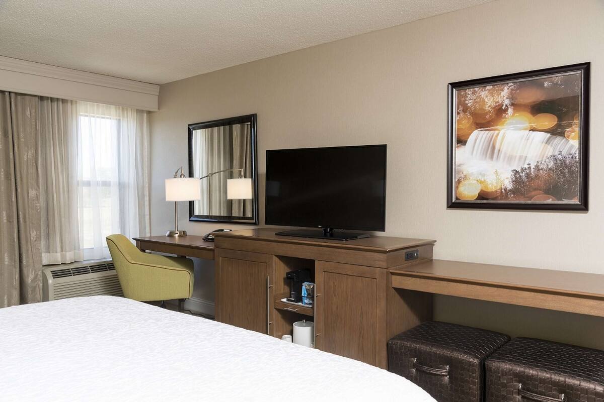 A hotel room with a bed, a desk with a flat-screen TV, a chair, a desk lamp, a framed picture of a waterfall, and a window with curtains.