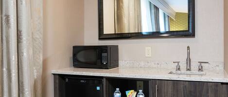 A bathroom with a microwave, sink, and a shelf with bottled water and snacks.