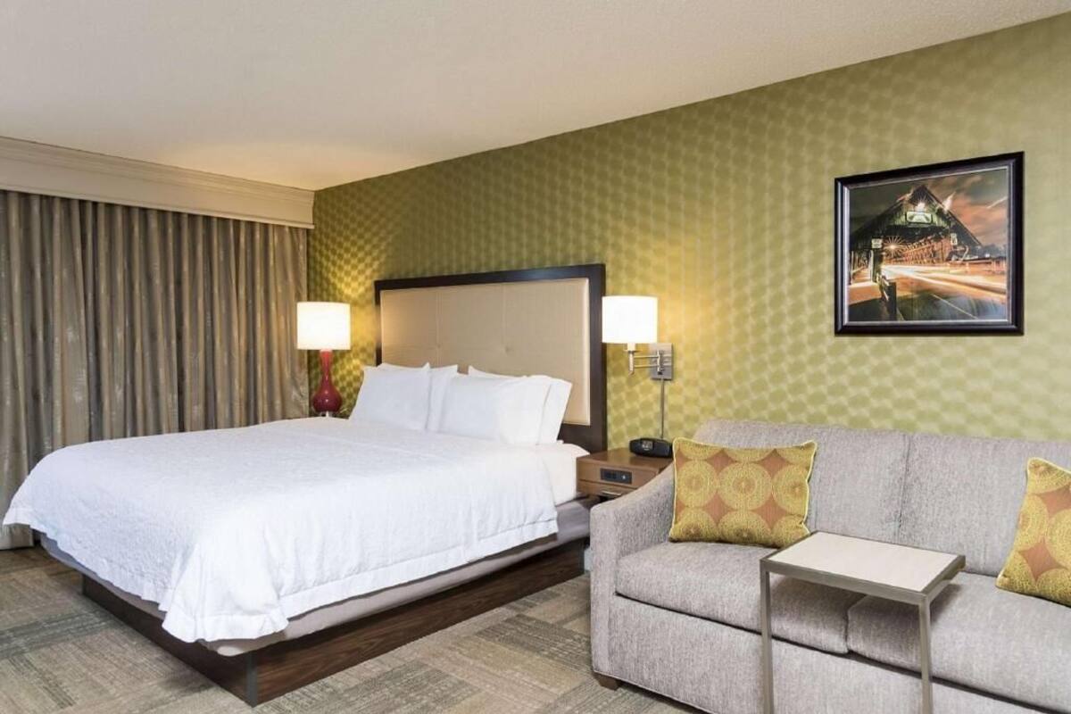 A hotel room with a bed, a sofa, a nightstand, a lamp, and a painting on the wall.