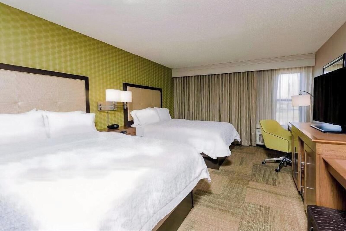 Una habitación de hotel con dos camas, un escritorio con una silla, un televisor y una ventana con cortinas.