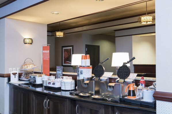 Un área de desayuno de hotel con un bufé, que incluye una máquina para hacer gofres, una cafetera y diversos alimentos.