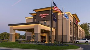 A Hampton Inn hotel with a modern design, featuring a prominent neon sign, American flags, and a well-maintained landscaped entrance.