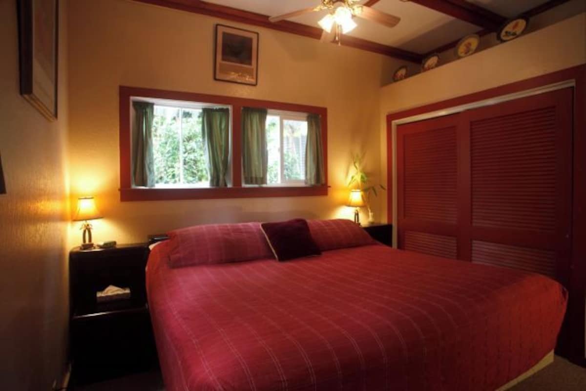 A bedroom with a large bed, two bedside tables with lamps, a window with curtains, and a ceiling fan.