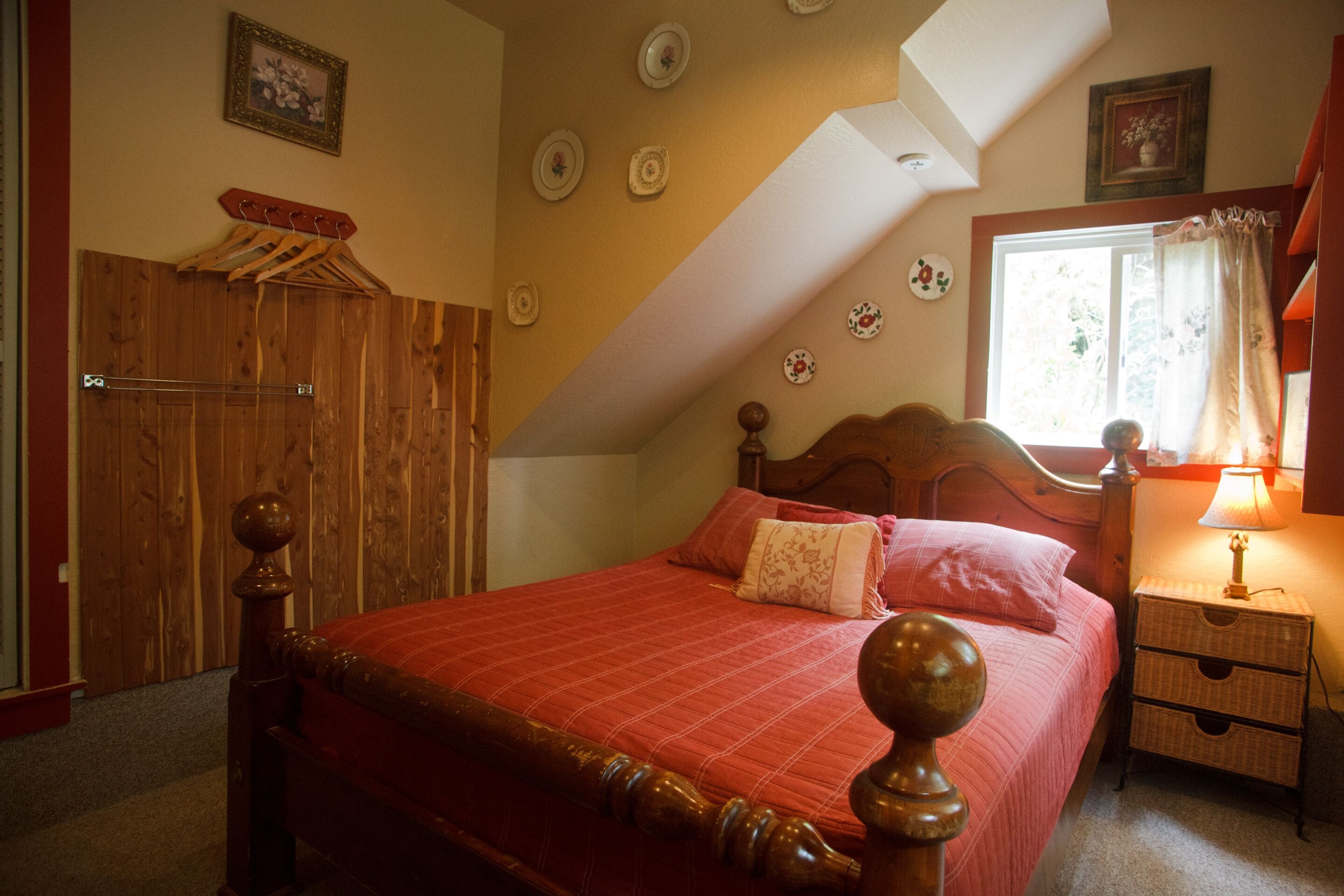 A bedroom with a wooden bed, a nightstand with a lamp, a window with curtains, and a wooden wardrobe.