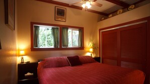 A bedroom with a large bed, two bedside lamps, a window with curtains, a ceiling fan, and a door with louvered shutters. - Volcano Hale (Volcano)