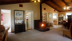 A cozy living room with a fireplace, wooden ceiling, and a ceiling fan. There is a dining area with a table and chairs, and a kitchen visible through an open door. - Volcano Hale (Volcano)