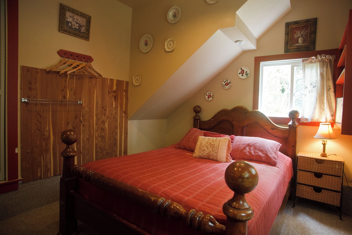 A bedroom with a wooden bed, a nightstand with a lamp, a window with curtains, and a wooden wardrobe.