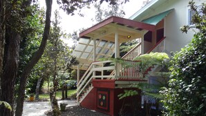 A house with a covered porch, a staircase, and a garden with trees and potted plants. - Volcano Hale (Volcano)