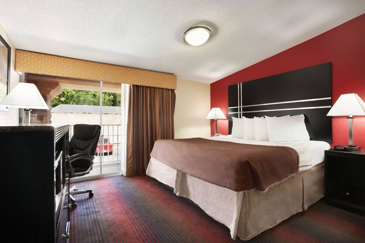 Une chambre d’hôtel comprenant un lit, deux lampes de chevet, un bureau avec une chaise, une fenêtre avec des rideaux et un mur d’accent de couleur rouge.