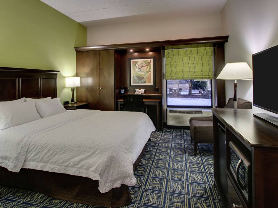 A hotel room with a large bed, a desk with a chair, a television, and a window with blinds.