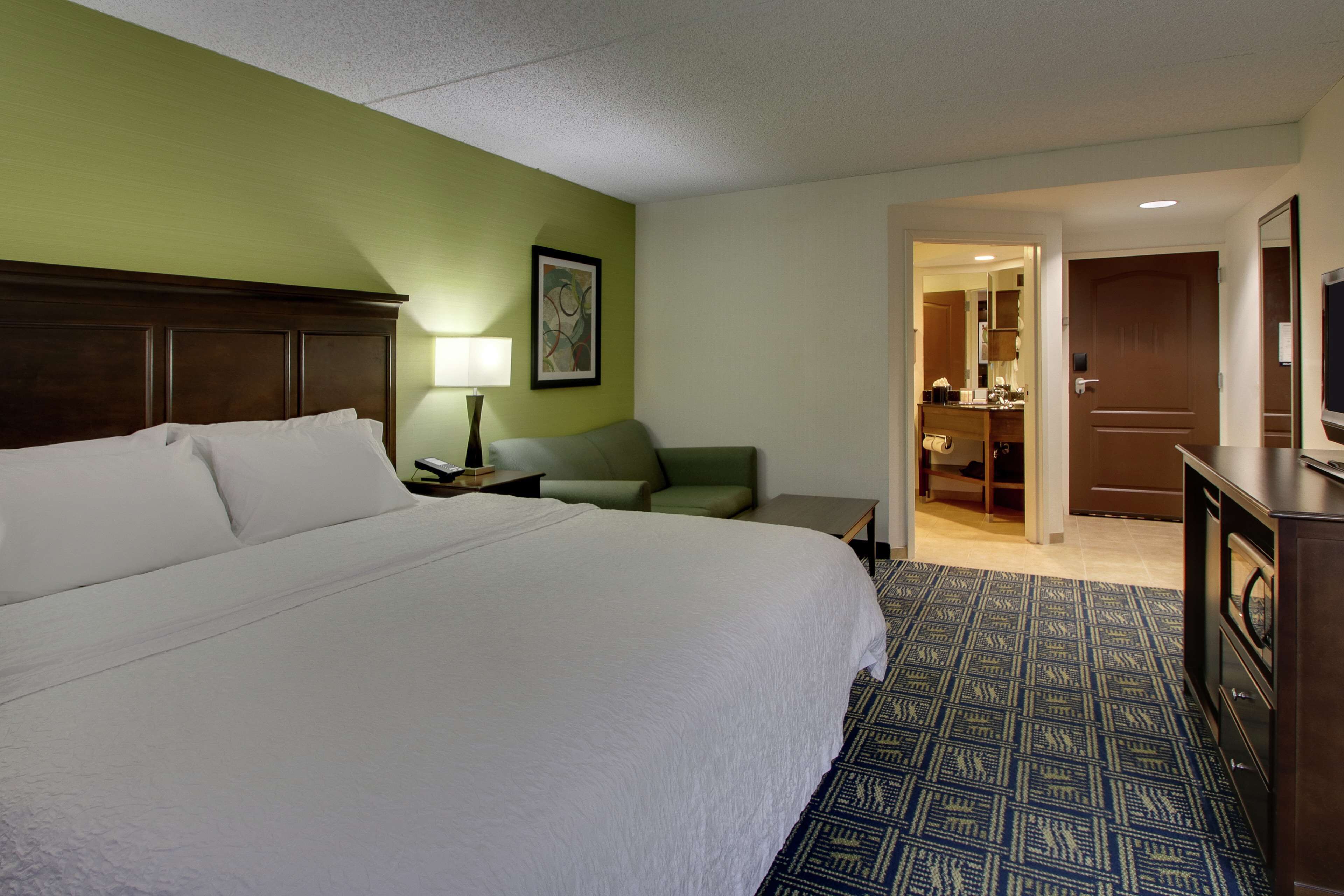 A hotel room with a large bed, a green wall, a patterned carpet, a desk with a lamp, and a door leading to a bathroom.