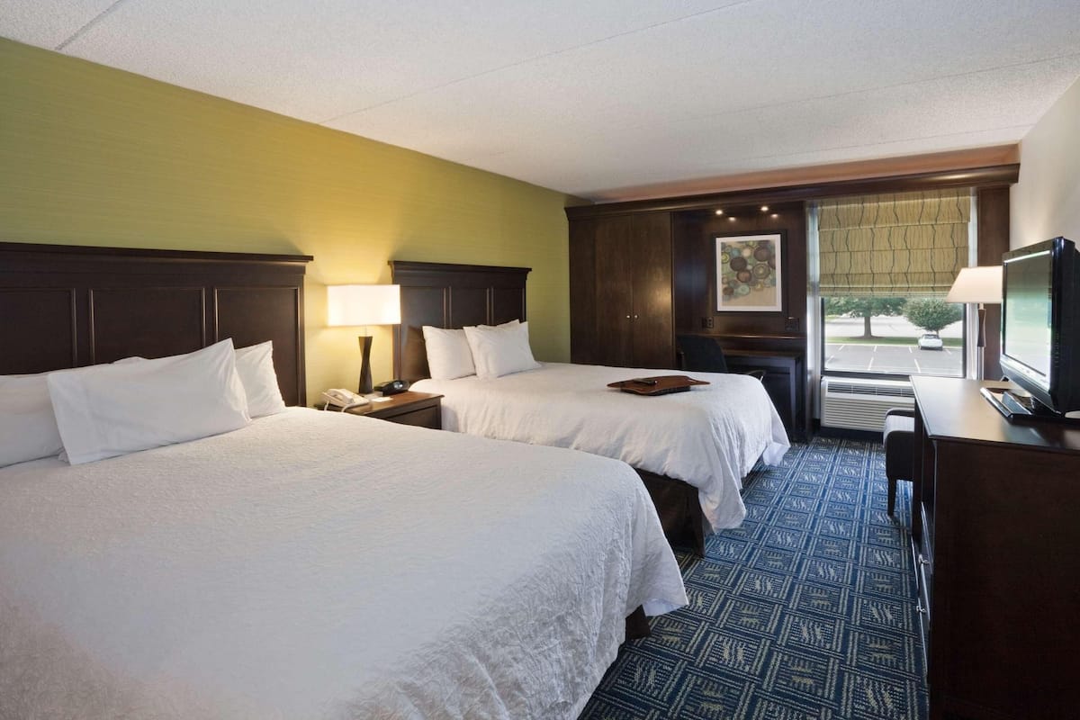 A hotel room with two beds, a desk, a TV, and a window with blinds.