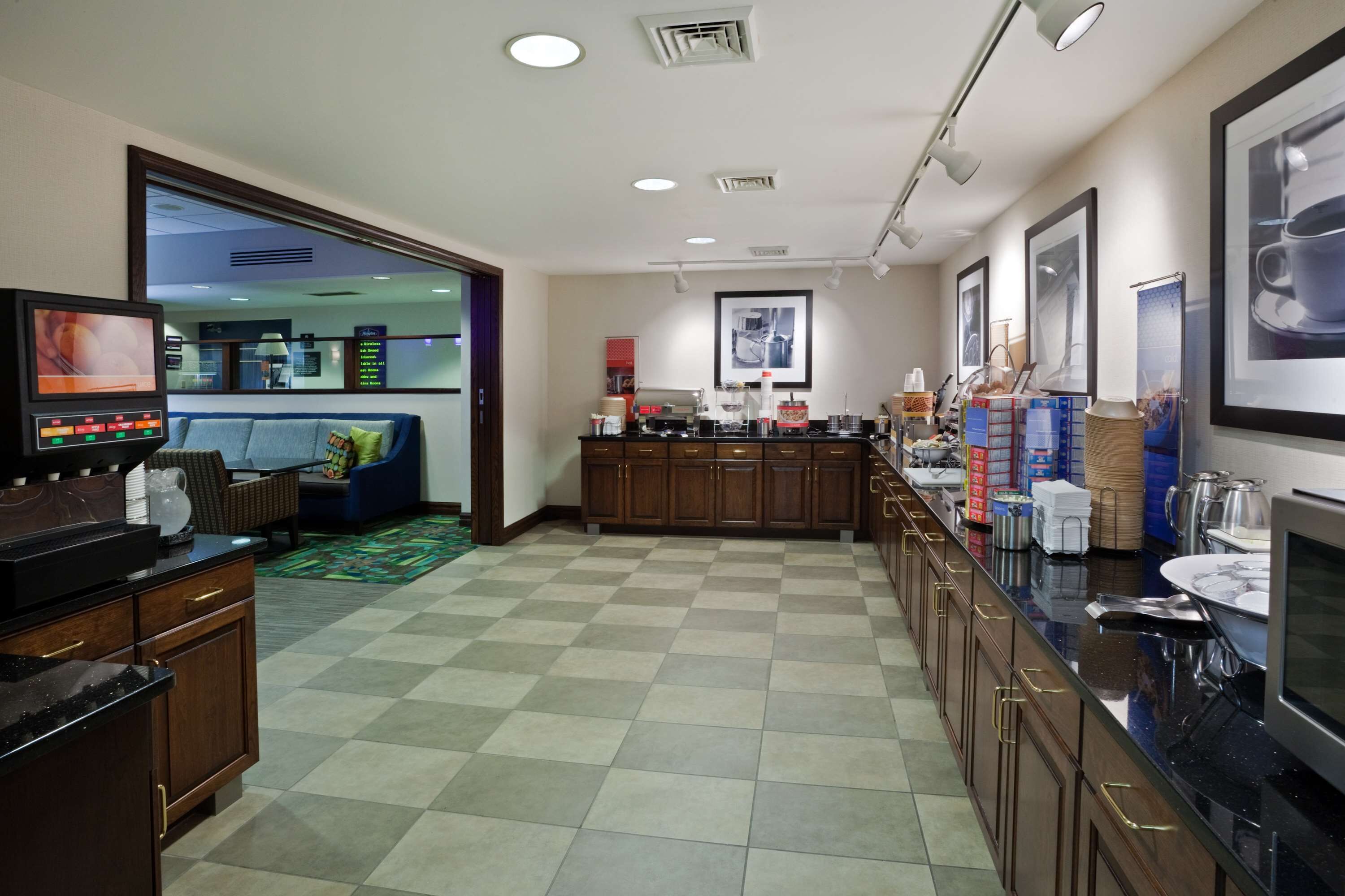 A hotel kitchen with a checkered floor, wooden cabinets, a coffee station, and framed pictures on the wall.