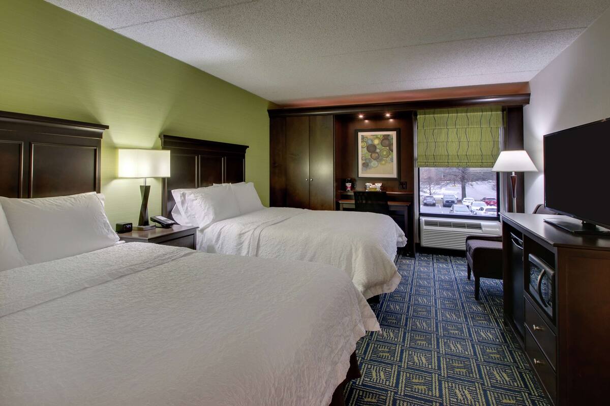 A hotel room with two beds, a desk with a lamp, a television, and a window with a view of parked cars.