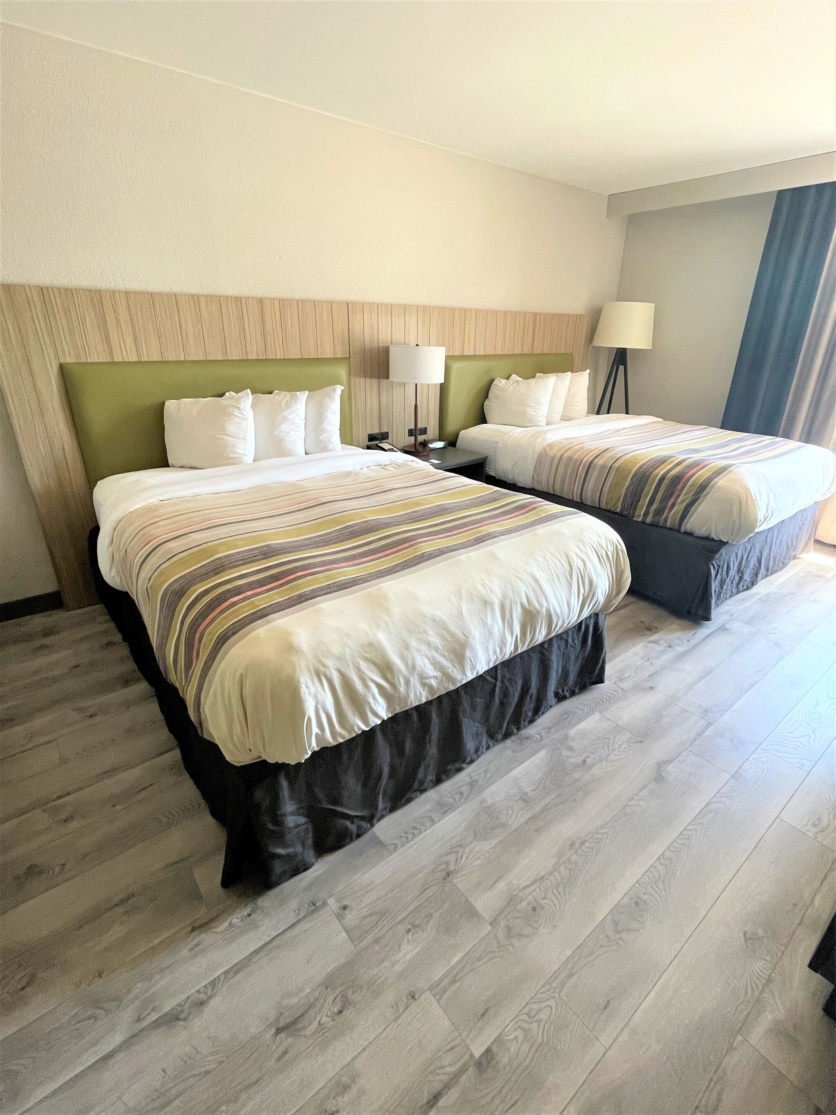 Habitación de hotel con dos camas, cabecero de madera, mesita de noche con lámpara y vista al exterior a través de una ventana.