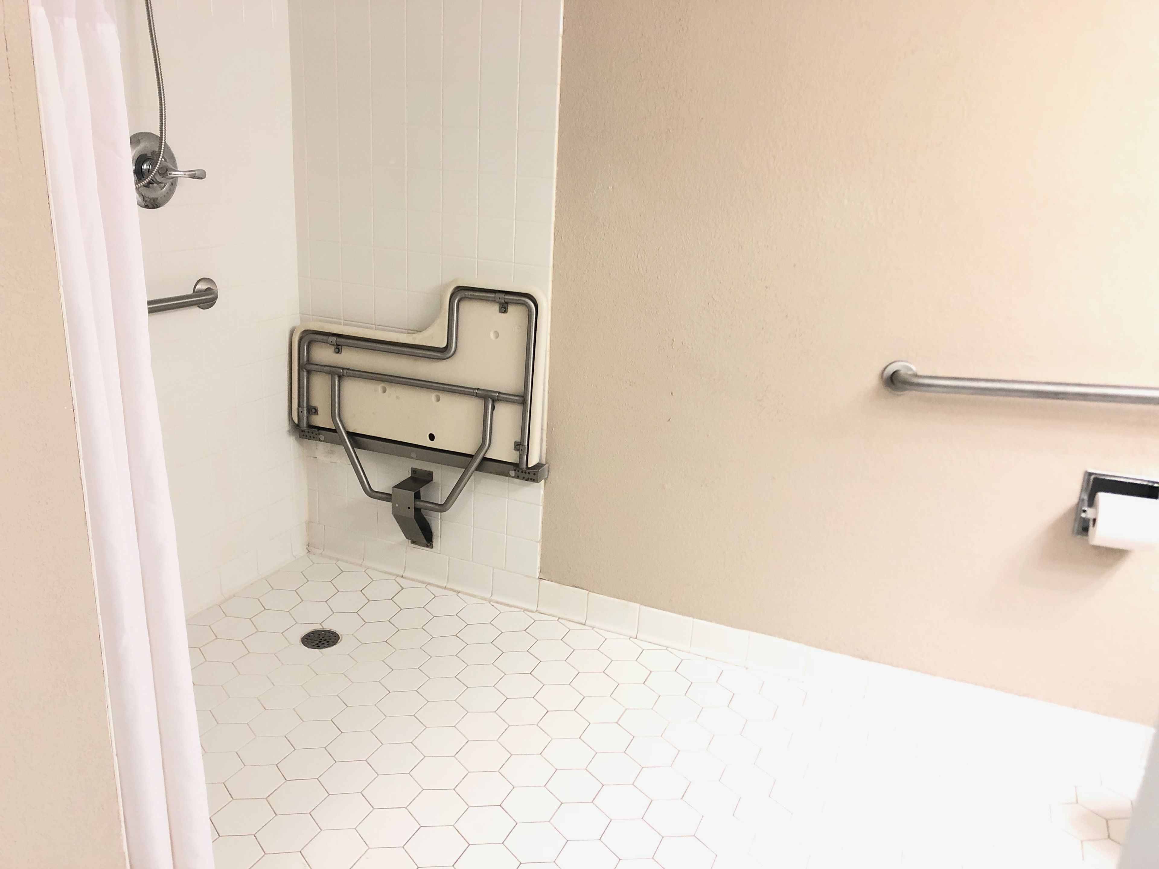 Un baño con piso de baldosas blancas, zona de ducha con barra de apoyo y un inodoro con barra de apoyo.