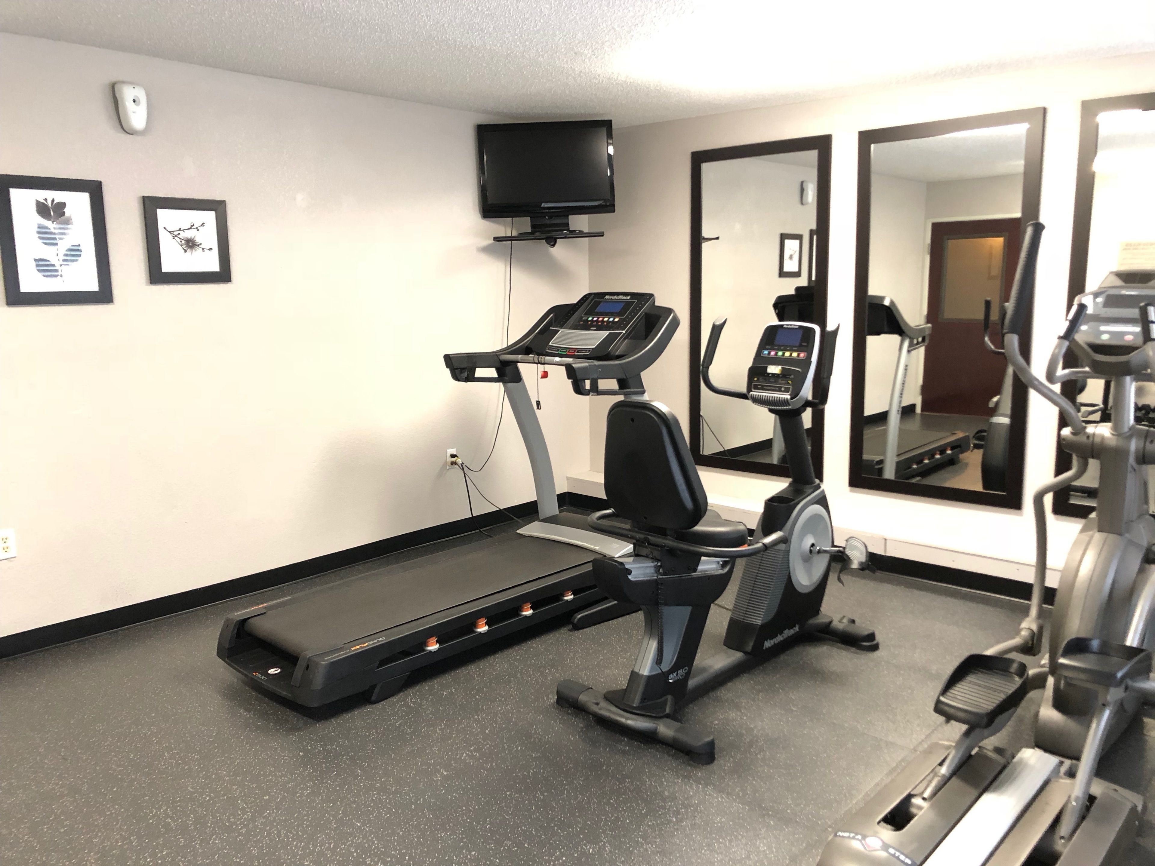 Un gimnasio con caminadora, elíptica y bicicleta estática, un televisor en la pared y obras de arte enmarcadas.