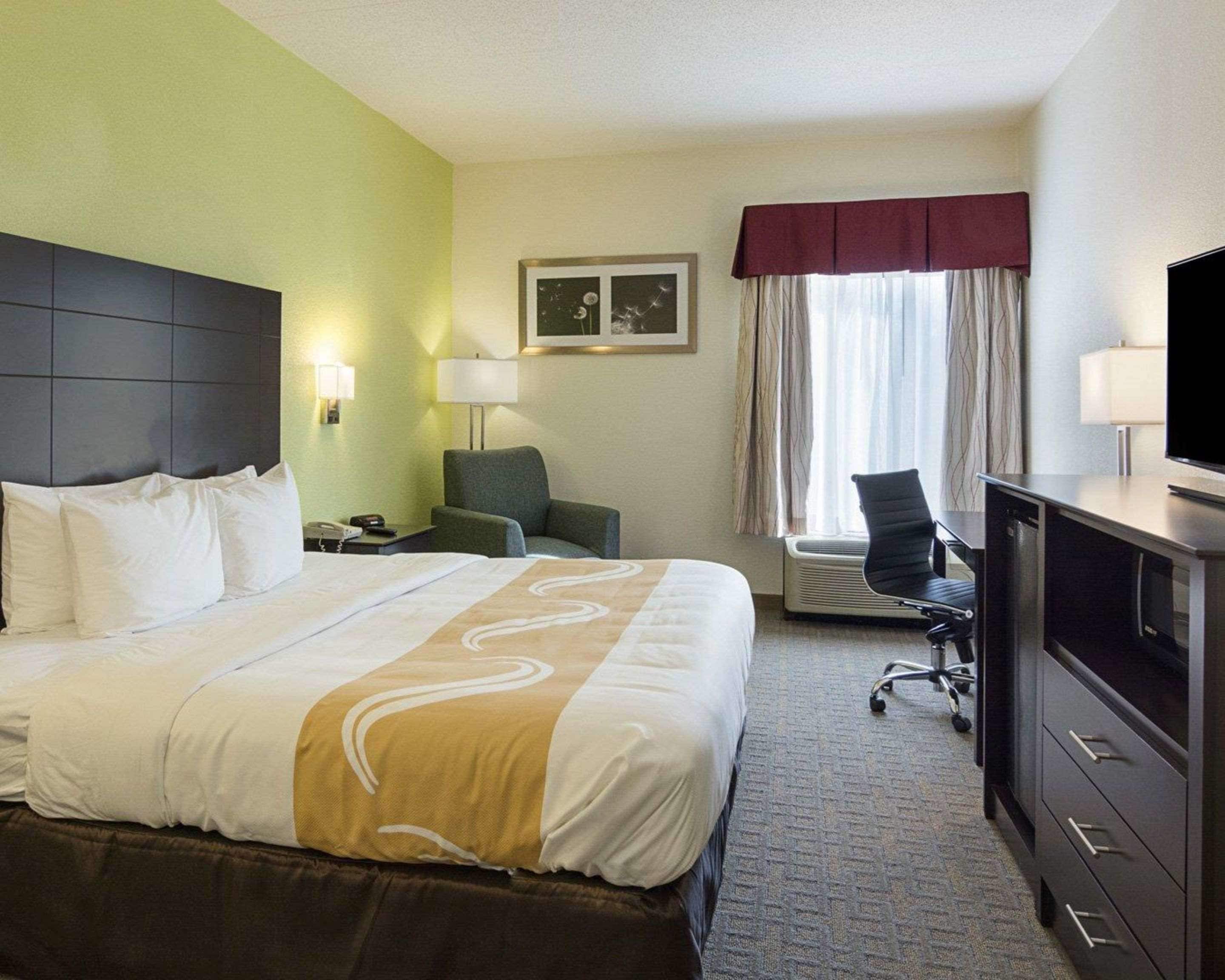 A hotel room with a large bed, a desk with a computer, a chair, a TV, and a window with curtains.