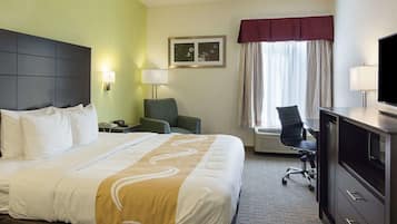 A hotel room with a large bed, a desk with a computer, a chair, a TV, and a window with curtains.