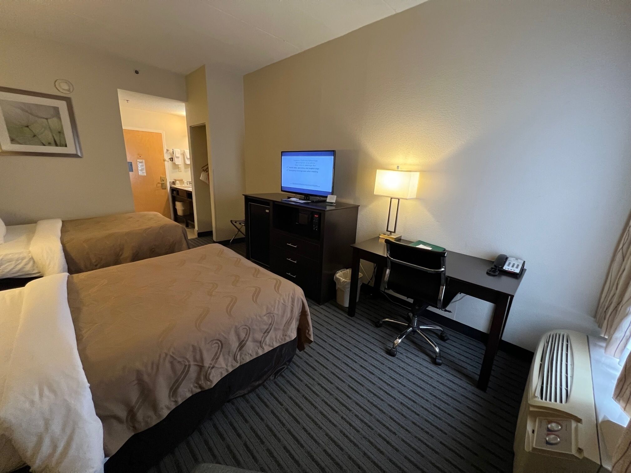 Quarto de hotel com duas camas, uma mesa com computador, abajur e telefone.