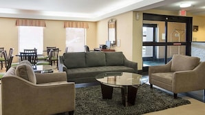 Un hall de hotel con un sofá, dos sillones, una mesa baja, una mesa de comedor con sillas y un ventanal con persianas.