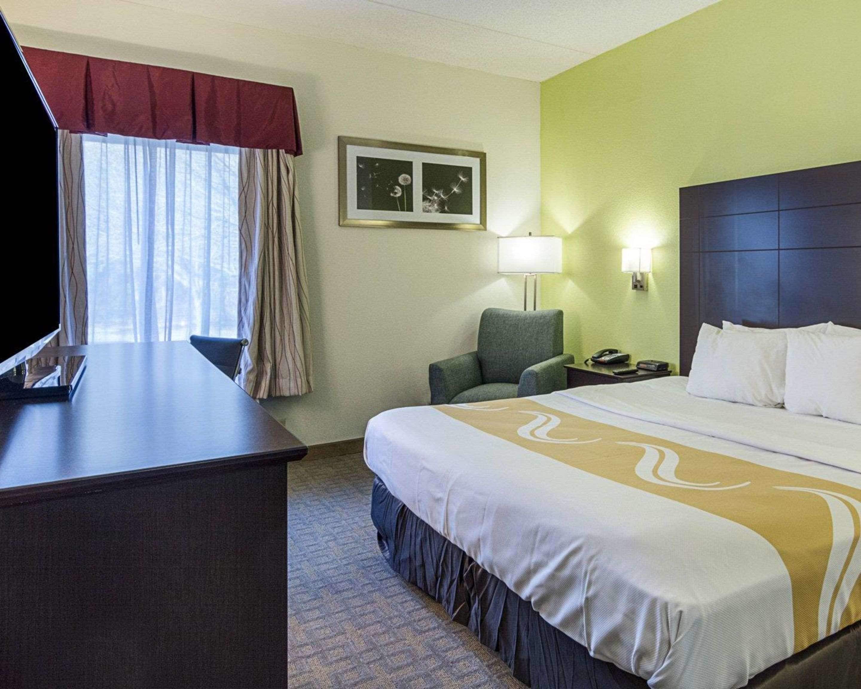A hotel room with a bed, a desk, a chair, a lamp, and a framed picture on the wall.