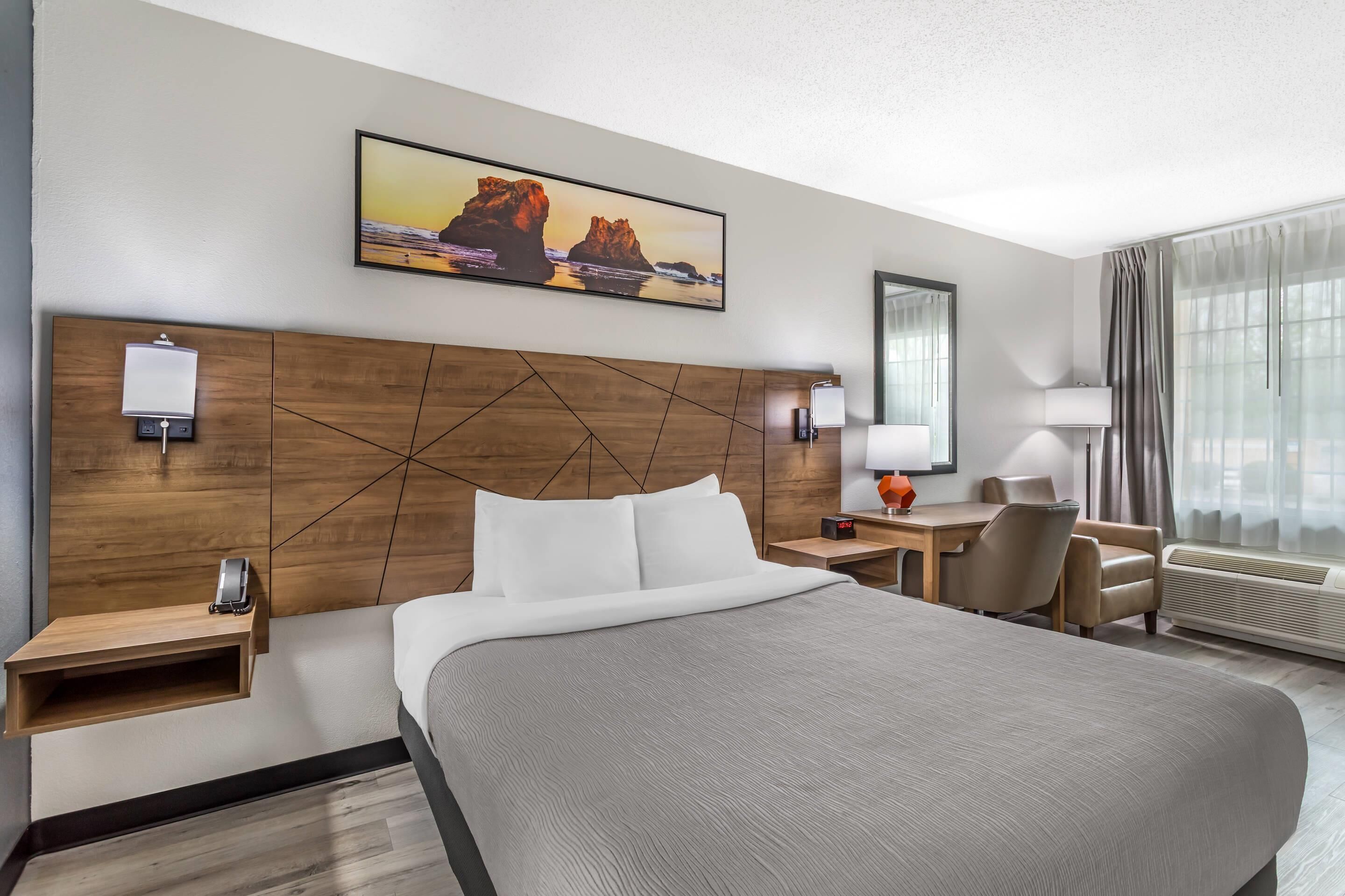 A hotel room with a bed, bedside tables, a desk, a chair, a lamp, a mirror, and a painting on the wall.