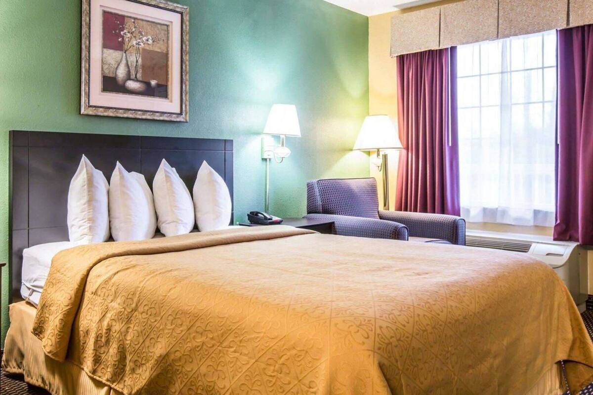 Une chambre d’hôtel avec un grand lit, un bureau, deux lampes, une chaise, un tableau encadré et une fenêtre avec des rideaux.