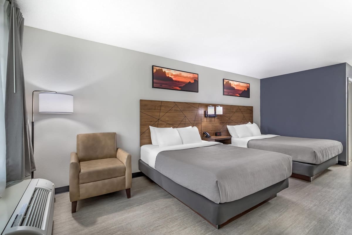 Une chambre d’hôtel avec deux lits, une chaise, un bureau avec une lampe et un climatiseur fixé au mur.