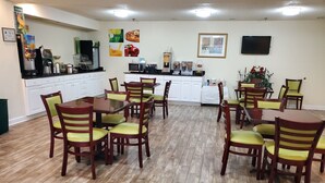 Un espace repas avec des tables et des chaises en bois, un coin cuisine équipé d’appareils électroménagers et un téléviseur fixé au mur.