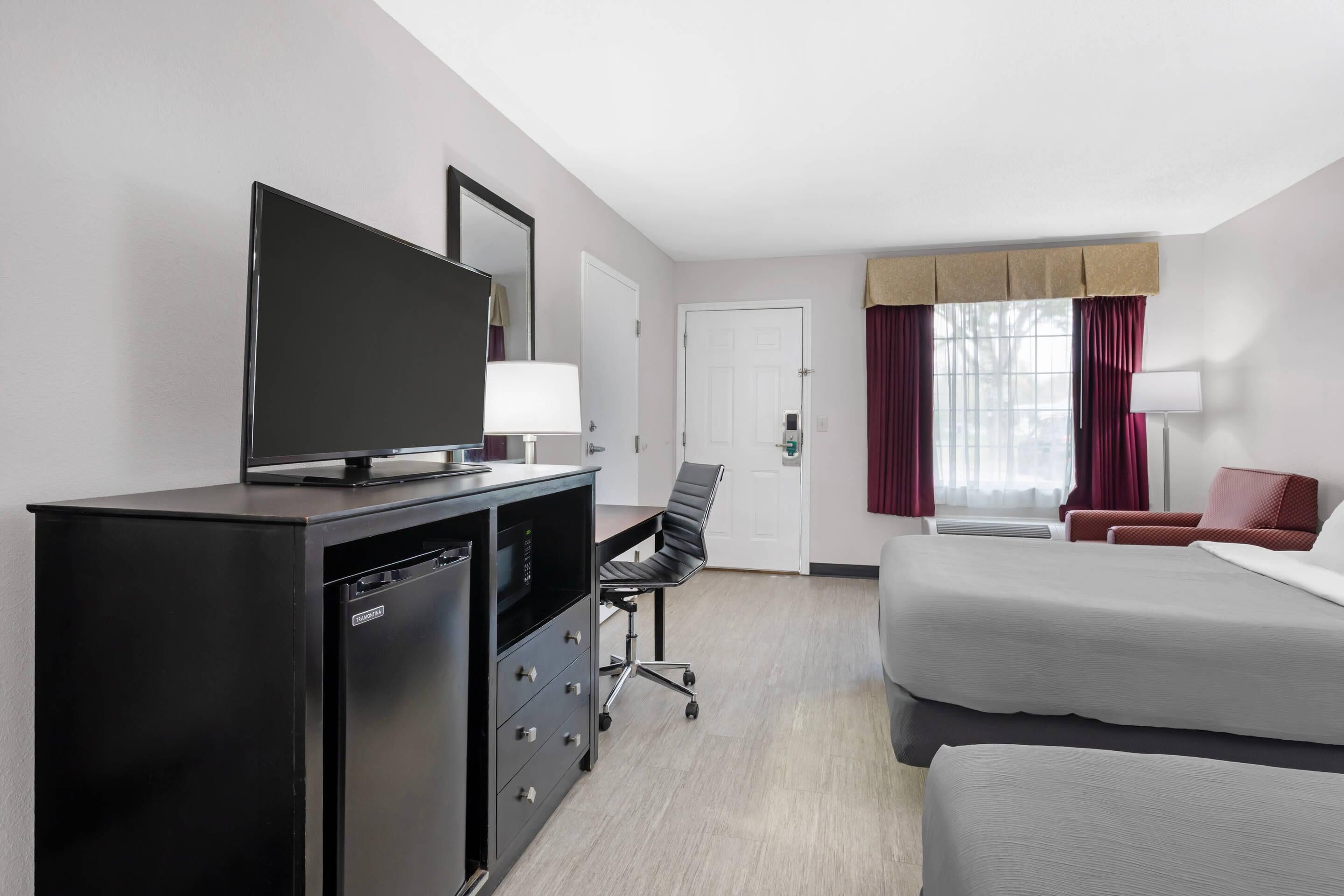 Habitación de hotel con cama, escritorio y silla, televisor, armario y ventana con cortinas.