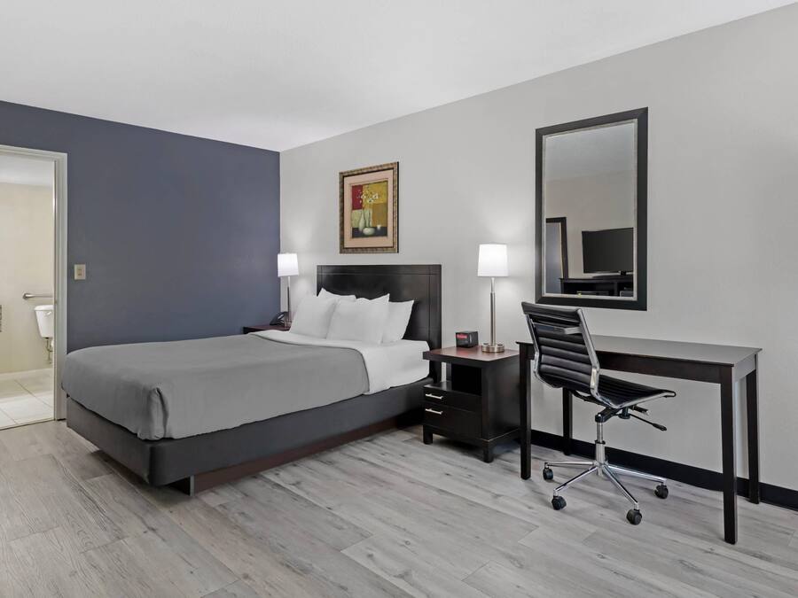 A hotel room with a bed, desk, chair, bedside lamps, and a painting on the wall.