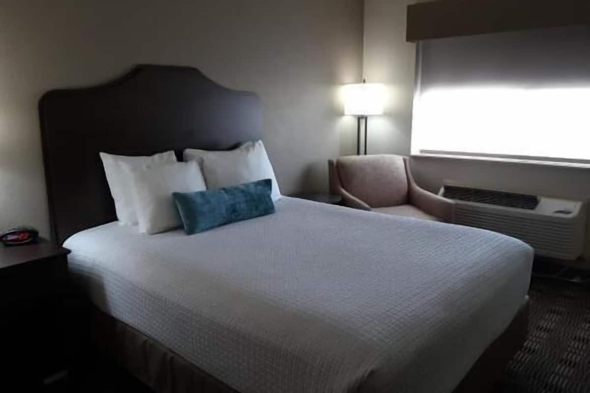 A hotel room with a large bed, a chair, a lamp, and an air conditioning unit.