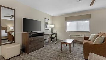 A hotel room with a flat-screen TV, a desk, a chair, a sofa, a coffee table, a mirror, a ceiling fan, and a window with blinds.