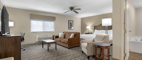 A hotel room with a bed, a sofa, a desk, a chair, a small table, a ceiling fan, a window with blinds, a lamp, and a bathroom with a bathtub.