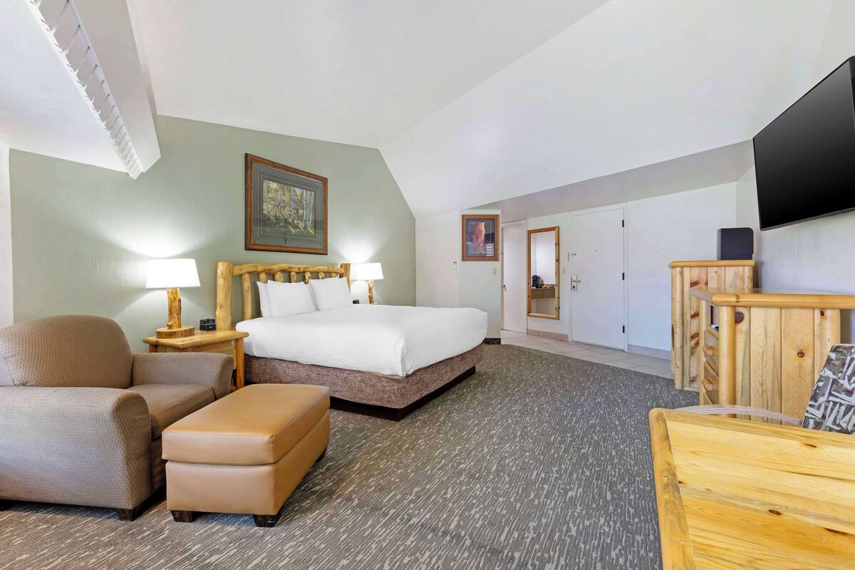Een hotelkamer met een bed, een stoel, een poef, een nachtkastje met lamp, een flatscreen televisie en een houten kast.