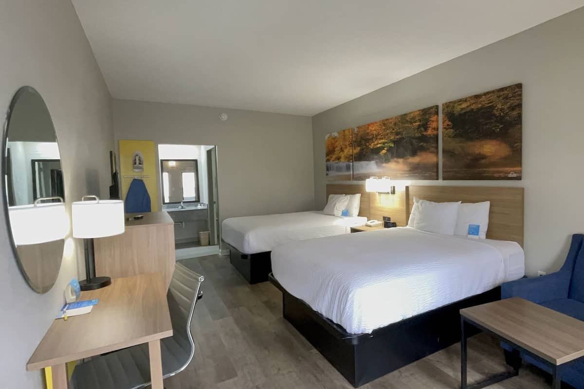 A hotel room with two beds, a desk, a chair, a mirror, and artwork on the wall.