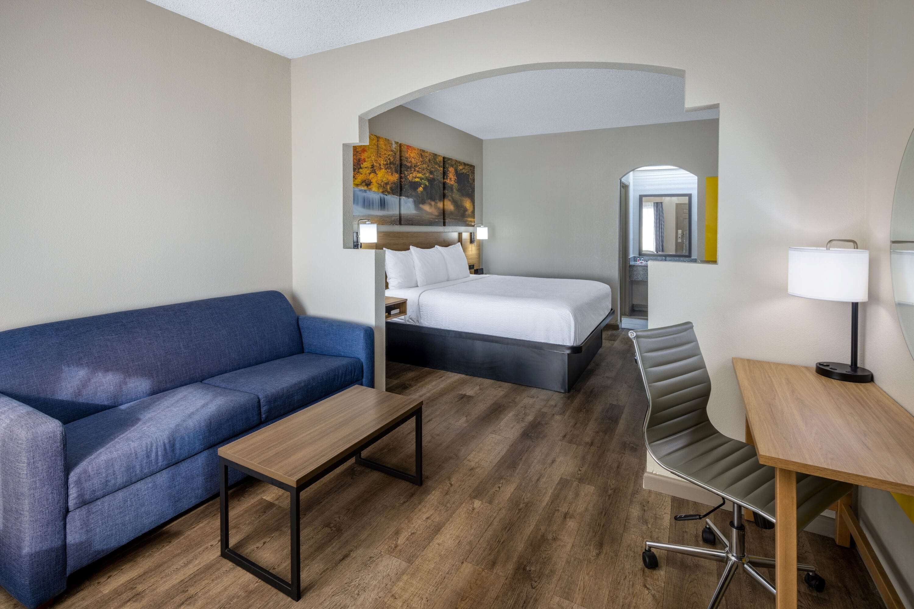 Una habitación de hotel con una cama, un sofá azul, una mesa de madera, un escritorio con una silla y un cuadro sobre la cama.