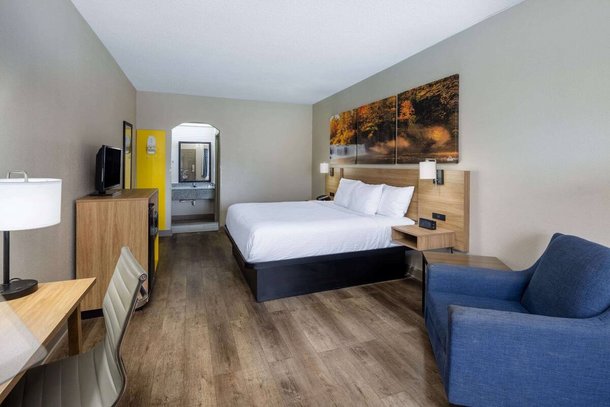 A hotel room with a bed, a desk, a chair, a TV, and a painting on the wall.