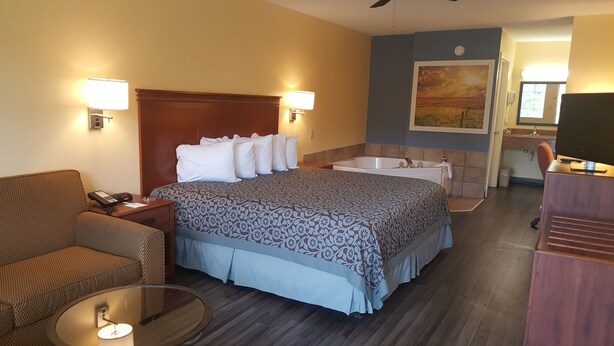 Ein Hotelzimmer mit einem Bett, einem Schreibtisch mit Telefon, einem Sessel, einem runden Tisch mit Lampe, einer Badewanne und einem Fernseher auf einem Schrank.