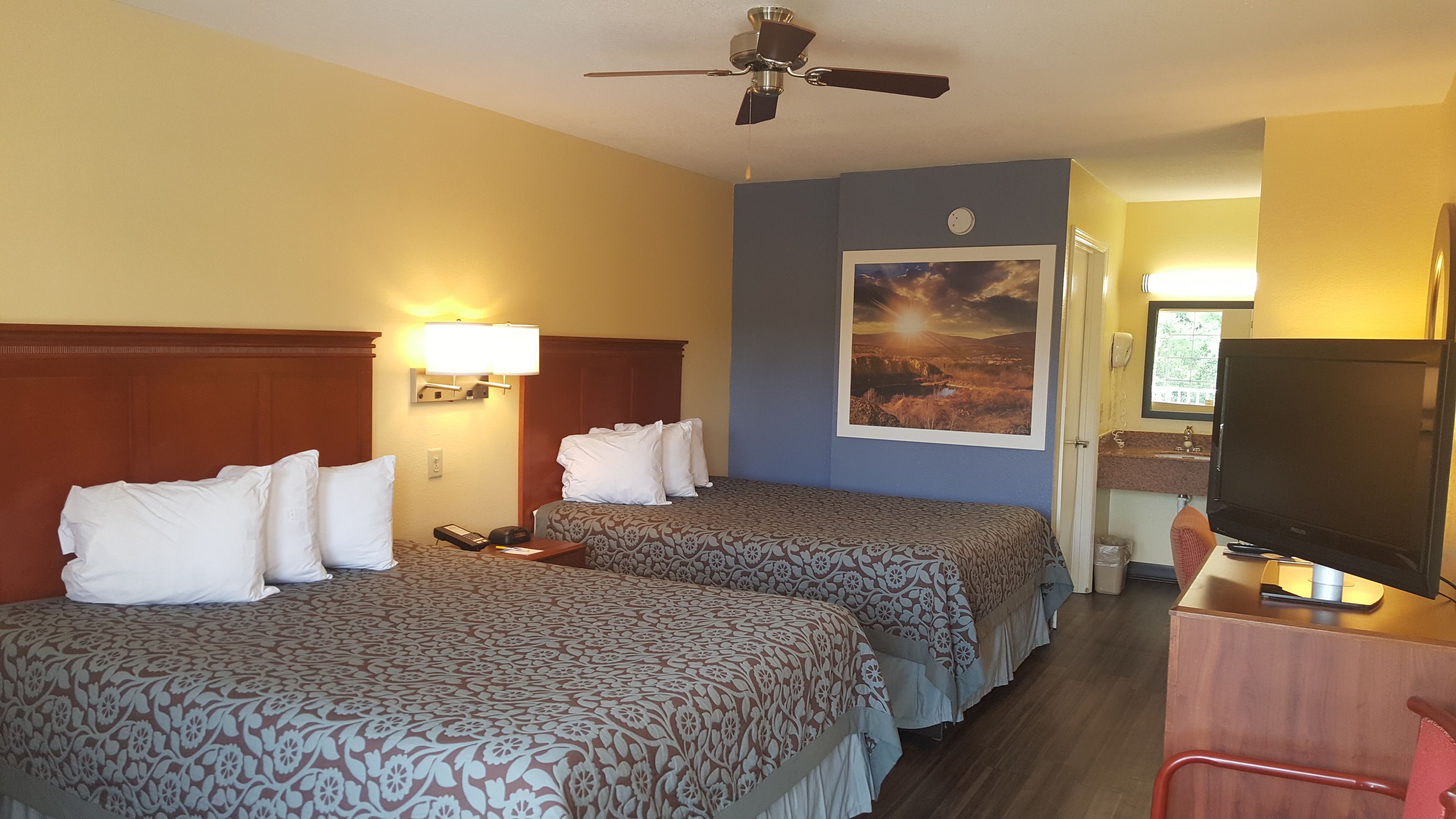 Quarto de hotel com duas camas, um ventilador de teto, uma luminária de parede, um quadro, uma escrivaninha com telefone e uma televisão.