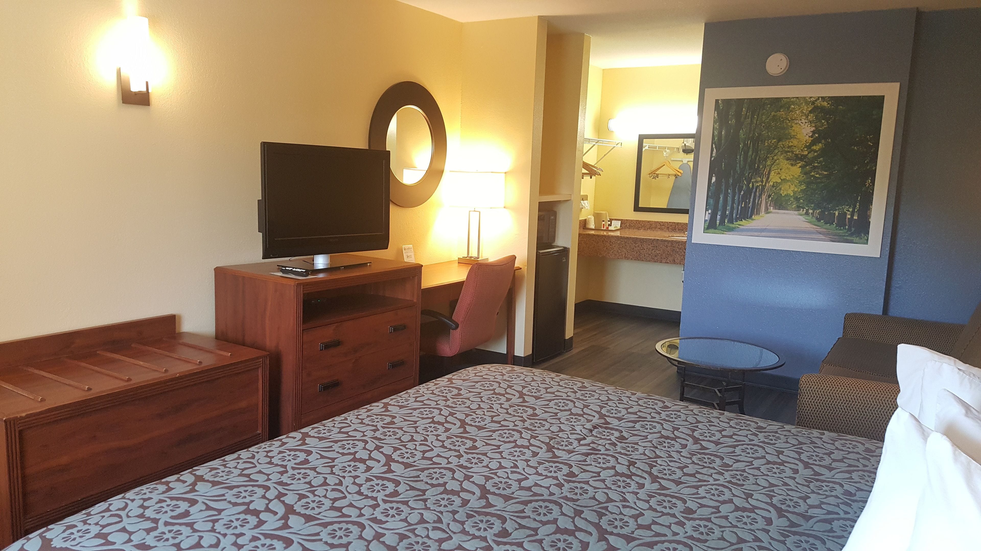 Ein Hotelzimmer mit Bett, Fernseher, Schreibtisch mit Stuhl, kleinem Tisch und Bad mit Spiegel.