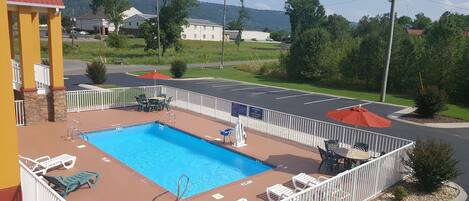 Una piscina con sillas de descanso y un área de terraza amurallada, rodeada por un estacionamiento y edificios al fondo.